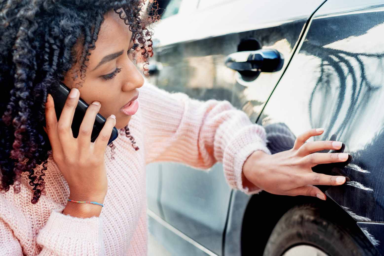 Young woman on phone inspects damage to her car.